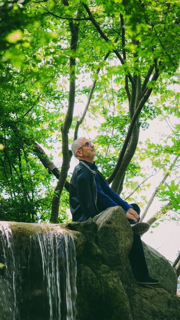 Senior man sitting peacefully by a small waterfall surrounded by lush green trees, enjoying nature's tranquility.